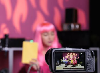 Influencer holding notebook showing product in front of camera while recording review using professional vlogging equipment. Asian content creator standing at desk filming vlog in broadcast studio